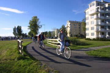 Flera cyklister på asfalterad väg vid vatten, gräsmattor och flerfamiljshus under klarblå himmel.