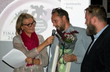 Charlotte Permell håller mikrofonen åt en man som håller blommor och ett pris, medan en annan man tittar på.