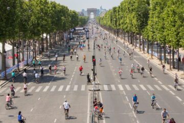 Många cyklister på en bred boulevard i Paris, med trädrader längs sidorna och Triumfbågen i fjärran.