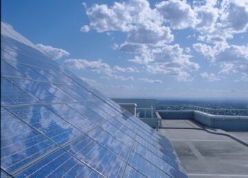 Solpaneler installerade i lutande rader på taket, reflekterar blå himmel och vita moln med utsikt över stadslandskap.