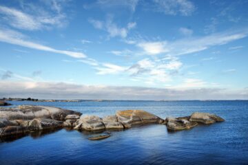 Slipade granitklippor i förgrunden, lugnt blått hav med små öar i horisonten och vid, molnprydd blå himmel.