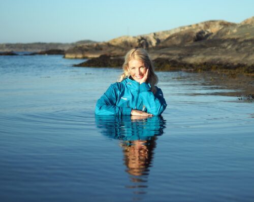 Kvinna i blå skaljacka står knädjupt i havet, ler med hand mot kind och syns speglad i vattnet.