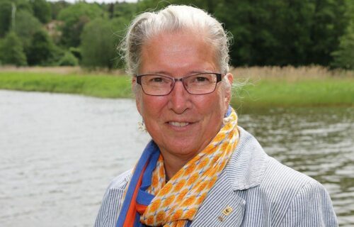 Porträttfoto av Eva Thörnelöf, en kvinna med grått hår, randig kavaj och orange-blå halsduk. Hon står vid vatten med grönska i bakgrunden.