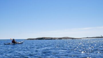 En person i livräddningsväst paddlar en kajak på stilla, solglittrande hav nära låga klippöar under klar himmel.