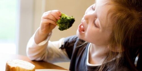 Liten flicka tar en tugga av en broccolibukett nära öppen mun, en skiva bröd syns på bordet.