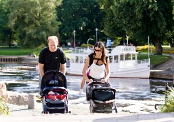Två vuxna promenerar med barnvagnar vid vattnet; en sovande bebis syns i vänstra vagnen. Text på båten: "SJÖSALA".