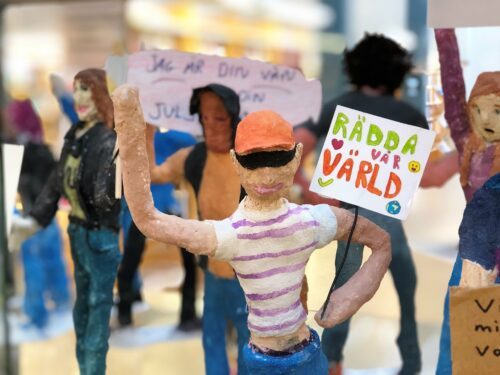 Handgjord demonstrantfigur håller skylt "RÄDDA vår VÄRLD" pappersmachéfigurer i demonstration.