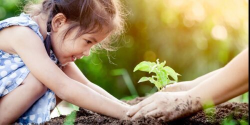 Flicka ler medan hon planterar en liten planta med hjälp av vuxna händer; båda har jordiga händer.