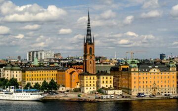 Riddarholmskyrkans spira reser sig över gula byggnader vid vattnet, med en vit passagerarbåt i förgrunden.