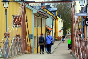 Par med barn och hund promenerar över en järnbron i Uppsala, mot färgglada trähus; en skylt visar "Järnbron".