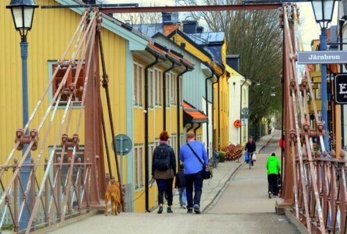 Kate-Elliott-shutterstock_570919564_1600x1080px, © Kate Elliott/Shutterstock Par med barn och hund promenerar över en järnbron i Uppsala, mot färgglada trähus; en skylt visar "Järnbron".