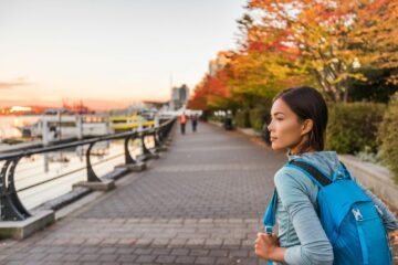 Ung kvinna med blå ryggsäck står vid kajpromenad, tittar framåt; höstfärgade träd och båtar i kvällsljus.
