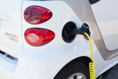 Bakpartiet på en vit elbil, röd baklampa. En gul spiralkabel är inkopplad i laddkontakt; text "electric drive" på karossen.