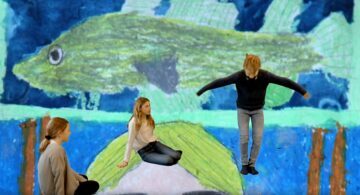Tre barn framför en stor, färgglad barnteckning av en grön fisk; två flickor sitter, en pojke hoppar med utsträckta armar.