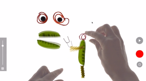 Två händer monterar ett lekfullt ansikte utav ärtskidor, spiralpasta och två runda ögon omgivna av röda cirklar.