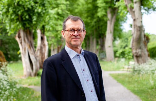 Porträttfoto av Mats Landén iklädd ljusblå skjorta och mörk kavaj. I bakgrunden syns en gångväg och gröna träd.