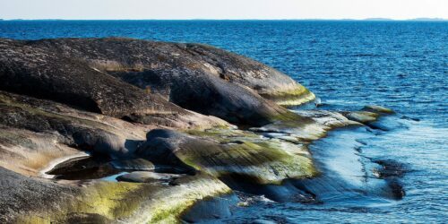 Släta granitklippor med gröna alger och vattenfyllda håligheter sluttar ner mot ett lugnt, blått innanhav.