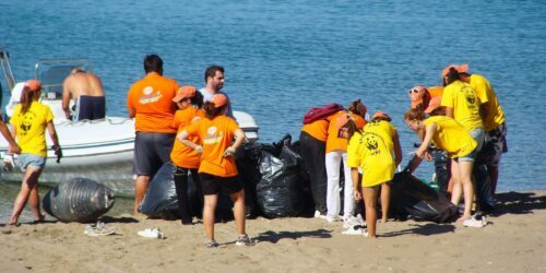 Frivilliga i gula och orange tröjor samlar svarta sopsäckar på en strand; de gula tröjorna visar tydligt den svarta WWF-loggan på ryggen. I bakgrunden syns även en vit båt.