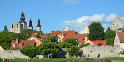 Bild av Almedalen, Gotland. Färgglada gamla hus och kyrktorn bakom lummig grönska vid en vattenspegel under klarblå himmel.