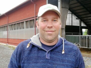 Porträttfoto av Holger van der Woude, i blå tröja och vit keps står framför en röd byggnad med fönster och öppning.