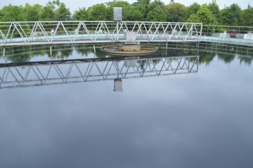 Cirkulär metallkonstruktion över stilla vatten och gröna träd i bakgrunden.