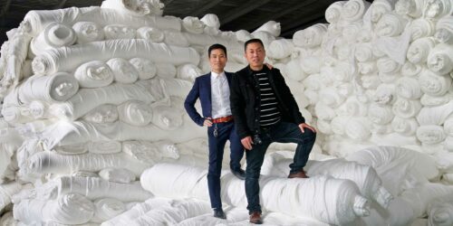 Two men stand against a backdrop of white colored textiles