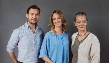 Tre står sida vid sida: man i blå och vit randig skjorta, kvinna i ljusblå blus, äldre kvinna i beige kofta, alla ler.