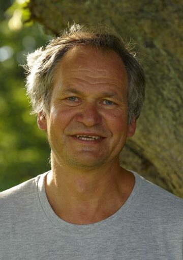 Porträttfoto av Peter Westman, leendes mot kameran. I bakgrunden syns en trädstam och grönska.