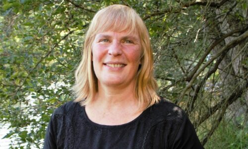 Porträttfoto Helene Grantz, axellångt blont hår och svart topp ler mot kameran, står framför flera träd.
