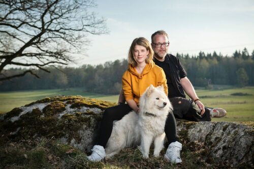 En kvinna och en man sitter utomhus på en sten med en stor vit hund mellan sig, med natur i bakgrunden.