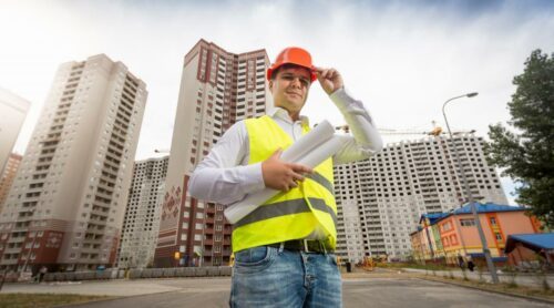 En byggnadsarbetare i gul varselväst och orange hjälm håller ritningar, med höga bostadshus i bakgrunden.