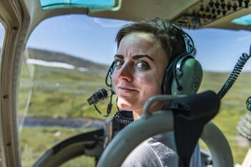 En kvinna med hörlurar i en helikopter vänder sig mot och tittar på betraktaren.