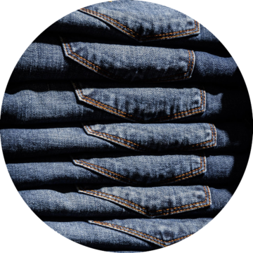 Rund bild: Flera hopvikt blå jeans med synliga bakfickor och orangea sömmar staplade i rad.