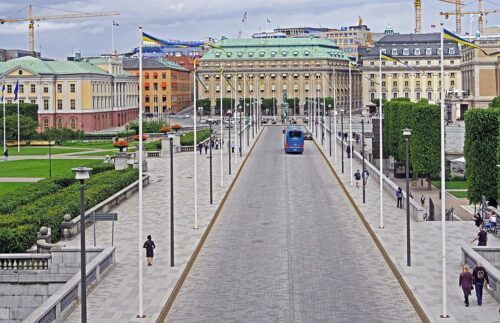 stockholm-3511345__480 Norrbron i Stockholm, kantad med flaggstänger med svenska vimplar. Bortom bron syns gamla byggnader med gröna tak. En blå buss syns på vägen.