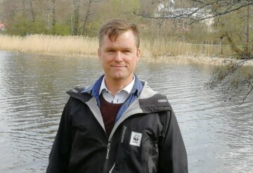 Porträttfoto av Gustaf Lind, iklädd svart jacka med WWF-logotyp och står vid en sjö med träd och vass i bakgrunden.