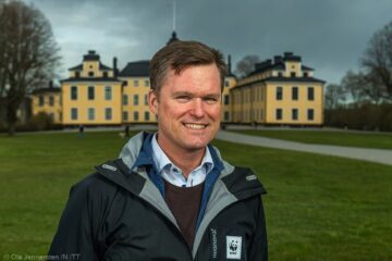 Porträttfoto av Gustaf Lind, i en mörk jacka med WWF-logotyp och blå skjorta. Han står utomhus framför Ulriksdals slott.