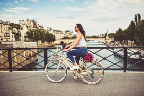 Kvinna cyklar på en cykel över en bro med ett flodlandskap och byggnader i bakgrunden, bär jeans och vitt linne.