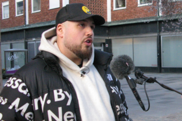 Anis Don Demina med skägg och keps intervjuas utomhus med mikrofon och bär svart jacka över vit hoodie.