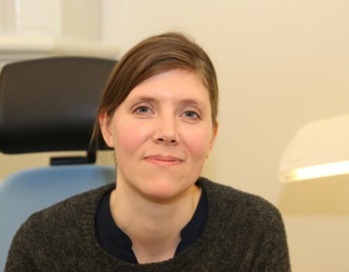 Porträttfoto av Anna Nolgren med uppsatt brunt hår, klädd i mörk tröja, sitter avslappnat med ett lätt leende.