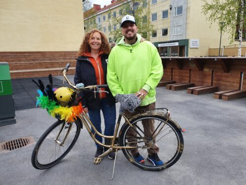 Anis Don Demina i Umeå, © Barbara Evaeus, WWF Två personer, en kvinna med rött hår och Anis Don Demina med grön huvtröja och keps, står bredvid en cykel med fjädrar längst fram vid cykelkorgen.