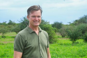GustafLindTanzaniaprio2023jpg Porträttfoto av Gustaf Lind, leendes mot kameran och står utomhus med träd och blå himmel bakom sig.