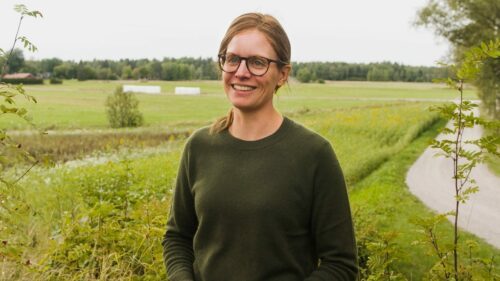 Louise, © Jimmie Petré En person i grön tröja står utomhus i grönskande fältlandskap med träd och öppna ängar i bakgrunden.