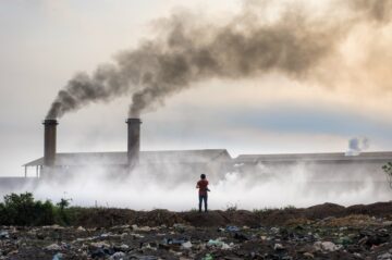 En person står på stenkust vid havet medan mörk rök stiger från en fabriksskorsten i bakgrunden.