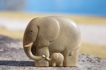 Grå elefantfigur i sten med en mindre grå elefant som står under den större. Figuren placeras på en slät yta, mjukt oskarp bakgrund.