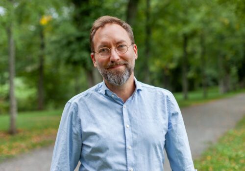 Porträttfoto av Ola Forshed i ljusblå skjorta. Ola står utomhus vid en stig, med gröna och suddiga träd och buskar i bakgrunden.