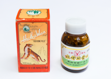 En orange kartong med texten "Huku Wan Tiger Bone Pills" och en illustration av en tiger bredvid en brun medicinflaska med kinesisk text och grön etikett.