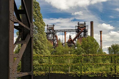 Utsikt mot en gammal industribyggnad med rostiga stålkonstruktioner och höga skorstenar, omgiven av tät grön vegetation och träd under en delvis molnig himmel. Bilden visar kontrasten mellan industriellt arv och frodig natur, med fokus på hållbar omställning och återbruk av industriområden.