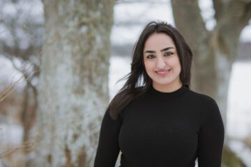 WSY 2023 Kimia 1 – foto BERNHARDINA HÖRNSTEIN, © BERNHARDINA HÖRNSTEIN Porträttbild på Kimia klädd i svart tröja står utomhus framför suddiga trädstammar med snö i bakgrunden.