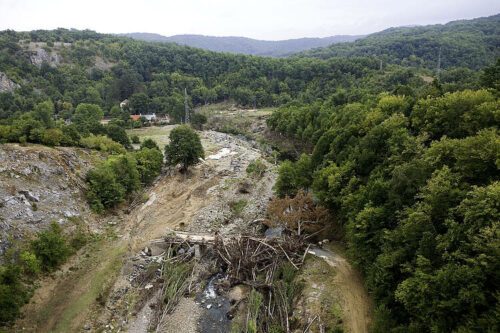 Översiktsbild över ett skogsklätt landskap där ett smalt vattendrag rinner genom en dal med tydliga spår av erosion och nedfallna träd efter ett kraftigt oväder. Bilden visar kontrasten mellan frodig skog och en skadad flodbädd, med fokus på naturkatastrofer, översvämningsskador och miljöpåverkan i ett bergigt område.