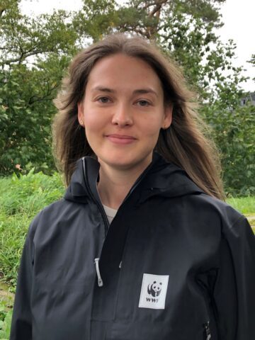 Sara Dreijer_WWF Porträttfoto av Sara Dreijer, med långt, brunt hår bär en svart jacka med WWF-logotyp, omgiven av grönska.